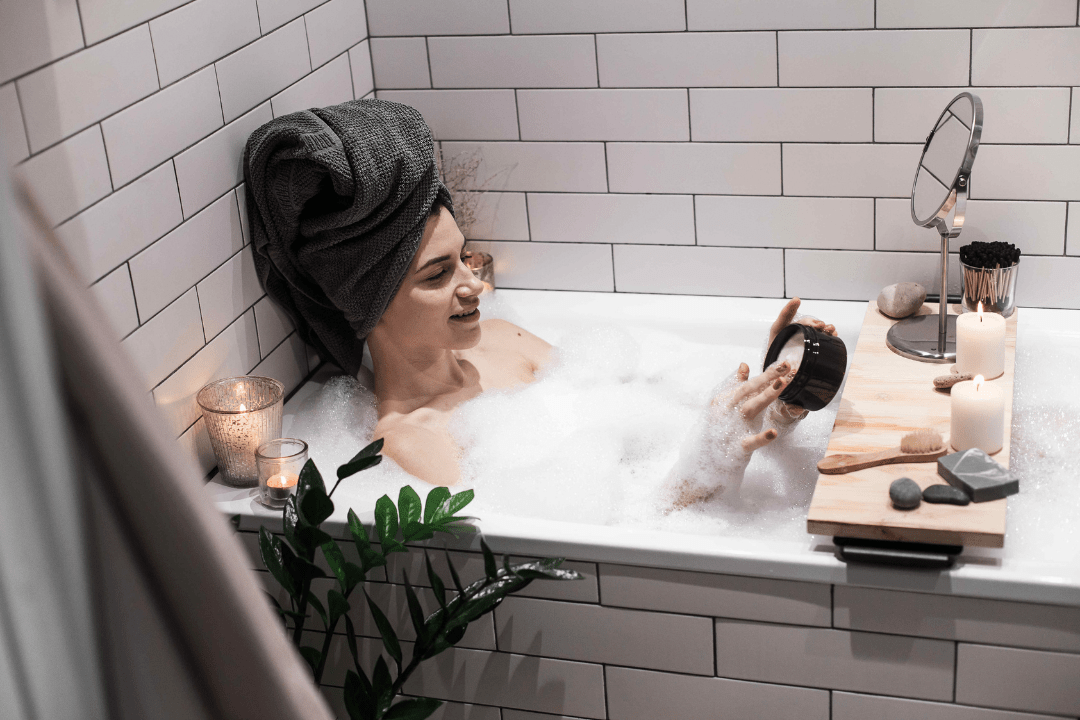 Richtig baden – So nutzen Sie die Vorteile eines Entspannungsbads optimal 1 Frau mit Handtuch über den Haaren in Badewanne mit Schaum sowie Pflegeprodukte griffbereit
